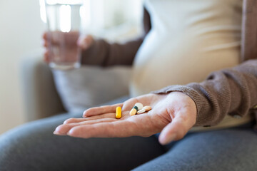 Obraz na płótnie Canvas Healthcare concept. Cropped view of pregnant woman take vitamin pill, holding cup with water in hands, sitting alone behind wooden table