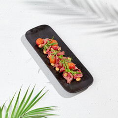 Roast beef with cherry tomatoes, ruccola and spicy sauce on black plate. Sliced roast beef on white concrete background. Italian antipasti in summer style with hard shadow and palm leaves.