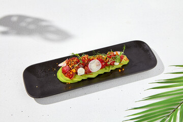 Modern food - tuna tartare on sliced avocado in black plate. Avocado starter with tuna and radish in asian style. Contemporary asian dish. Healthy food with avo and marinated fish on white background.