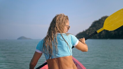Long curly haired blonde woman in swimsuit rows plastic kayak along azure ocean water at exotic resort close up backside back view. Traveling to tropical countries. Girl is sailing on kayak in ocean.