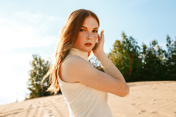 Young redhead woman in dress touching hair on sand in forest and looking at camera 