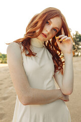 Fototapeta na wymiar Portrait of young gentle woman with long ginger hair touching head and looking at camera on sand in nature 