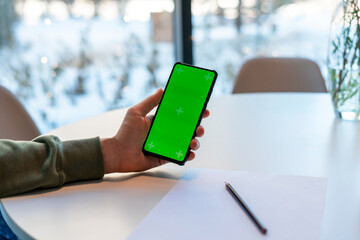 Man Holding Smartphone with Chroma Key Screen in cafe at white table, makes notes with pencil on paper , green screen man's hands mock up