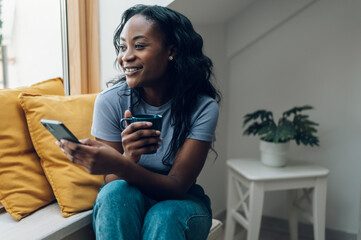 Obraz premium African american woman sitting on windowsill with a cup of coffee and a smartphone