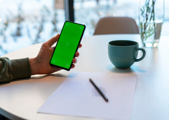 Man Holding Smartphone with Chroma Key Screen in cafe at white table, makes notes with pencil on...