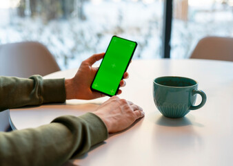 Man Holding Smartphone with Chroma Key Screen in cafe at white table, drinking coffee green screen man's hands, business mock up