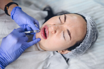 Cosmetologist doing painful lip augmentation procedure with hyaluronic acid. The beautician pierces...