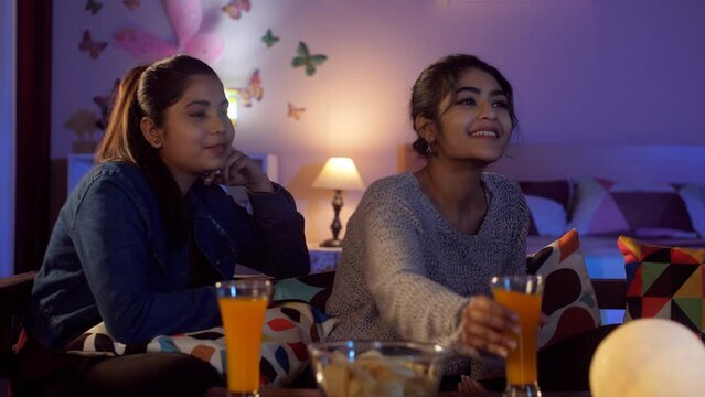 A pretty young college student changing television channels using a remote control - an electronic device channel surfing entertainment. Two best friends eating snacks and drinking juice while we...