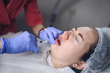 Fototapeta premium Cosmetologist doing painful lip augmentation procedure with hyaluronic acid. The beautician pierces lips by needle. Woman suffering subcutaneous injection to increase lips shape with dermal filler.