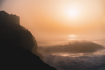Silhouette of a man sitting on a cliff watching a giant wave breaking with the sunset in the background.