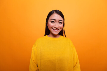 Beautiful happy asian woman smiling heartily while wearing yellow sweater on orange background. Gorgeous attractive joyful person wearing colorful blouse while smiling at camera.