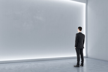 Man standing in modern exhibition hall interior with illuminated white mock up wall and concrete floor. Gallery concept.