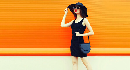 Portrait of beautiful young woman with handbag wearing a black summer hat and dress on orange...