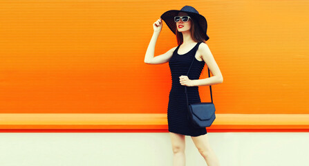 Portrait of beautiful young woman with handbag wearing a black summer hat and dress on orange background