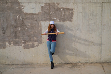 Obraz na płótnie Canvas young and beautiful redhead woman is happy with baseball cap, jacket, baseball bat and jeans, she is posing in front of grey concrete background. Concept sport and recreations.