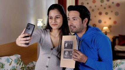 A young man and his beautiful pregnant wife on a video call while using a mobile. A cheerful guy showing an ultrasound report while chatting online - new parents, motherhood, positive news