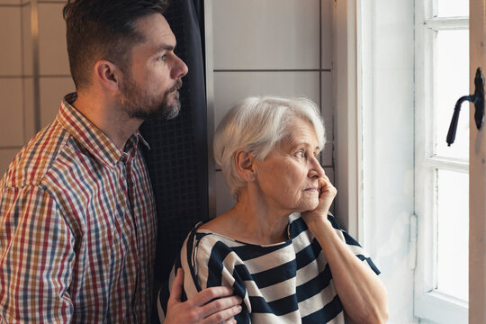 Caucasian Elderly Pensioner Mother And Her Middle-aged Son Worried Looking Through The Window At A Serious Accident Outside. High Quality Photo