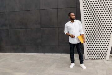 Portrait of confident IT person in formal outfit standing near the office, holding computer, looking to the camera. Stylish dressed african american male freelancer isolated at grey background, posing