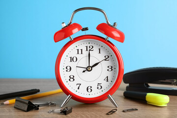 Red alarm clock and different stationery on wooden table against light blue background. School time