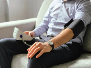 Fototapeta na wymiar Man is checking blood pressure at home. Sitting on a grey sofa. Monitoring of Seasonal health issues or stress at work Hypertension or hypertension seasonal disorder and problems Taking care of health
