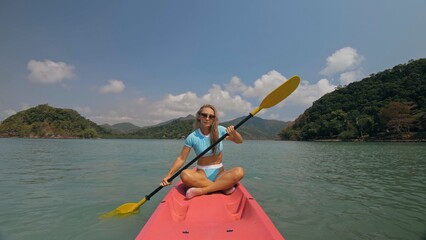 Attractive sportive blonde woman with sunglasses rows pink plastic canoe along sea water against...