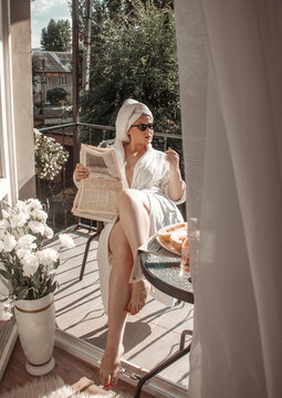young girl in a white bathrobe and towel on head is sitting near flowers in black sunglasses and newspaper in hand on green trees background on french balcony at morning. lifestyle concept, free space