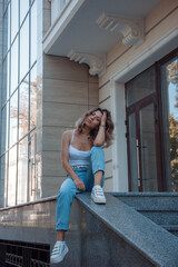 young curly brunette girl in white top and blue jeans is sitting casual with hand near head and looking straight near modern glass building background. lifestyle concept, free space