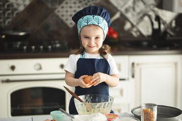 A cute little girl in an apron and a Chef's hat breaks an egg into a dish, looking at the camera and smiling while cooking.