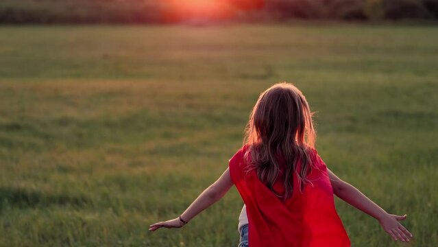 Girl Plays Superhero. Happy girl are playing superhero. Kid run across green field in red cloak at sunset time. Pretty child superhero hero in red cloak in nature. Girl power concept.