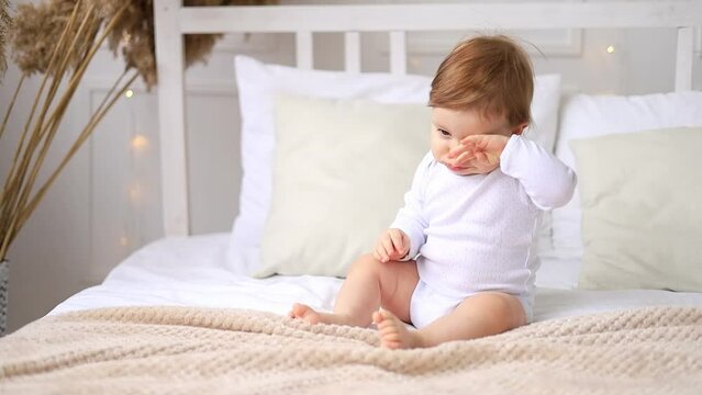 little baby girl rubs her eyes with her hand and wants to sleep on a white cotton bed at home in a bright room