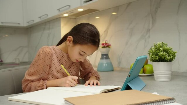 Adorable preteen school girl writing in copybook, raising hand to answer question of tutor while studying via online educational platform, using digital tablet at home. E-learning concept. Moving shot