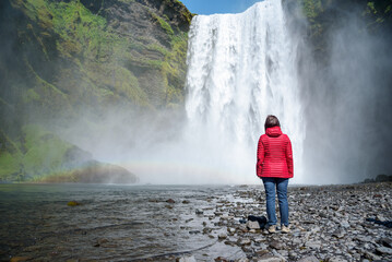 Woman standing alone in front of a mighty waterfall on a sunny summer day. Skogafoss waterfall,...