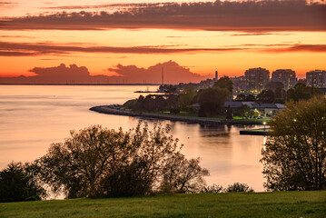 Fiery sunset over a city on the shore of a lake. An island dotted with tall wind turbines is...