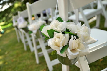 White Chairs Adorned with White Fabric Rose Bouquets Await Guests at a Garden Wedding