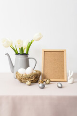Blank photo frame, nest with Easter eggs, rabbit and tulips on table near light wall
