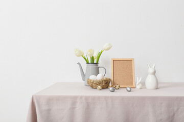 Blank photo frame, nest with Easter eggs, rabbits and tulips on table near light wall