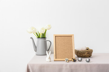 Blank photo frame, nest with Easter eggs, tulips and rabbit on table near light wall