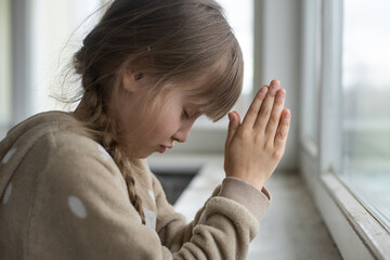 Praying little girl near window - obrazy, fototapety, plakaty