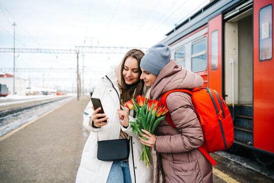 two girls on the station platform with flowers happily looking into a smartphone