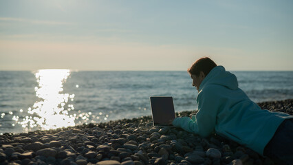 Caucasian woman lying on her stomach on a pebble beach and typing on a laptop. Remote work freelancer. © Михаил Решетников