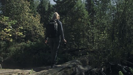 Obraz premium Traveler photographing scenic view in forest river. Wood bridge fallen tree. One caucasian woman shooting nice magic look. Girl take photo video on camera. Photographer travel with backpack. Outdoor.
