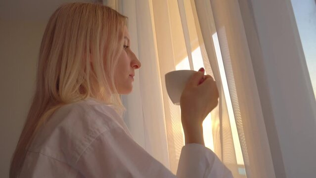 A young beautiful blonde in a white shirt looks out the window, drinks a hot drink of tea or coffee, the rays of the setting or rising sun fall on her face. Tenderness and softness.