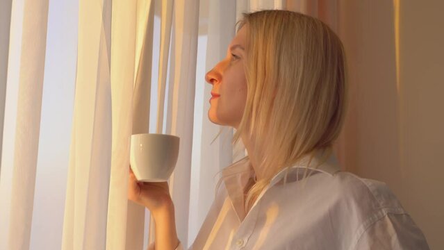 A young beautiful blonde in a white shirt looks out the window, drinks a hot drink of tea or coffee, the rays of the setting or rising sun fall on her face. Tenderness and softness.
