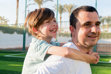 Side view of smiling father carrying cheerful boy on back while spending time together at football field