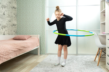A child spins a hoop at home in the living room. Little girl with a dancing black dress twists a hoop at home in the living room © Natallia