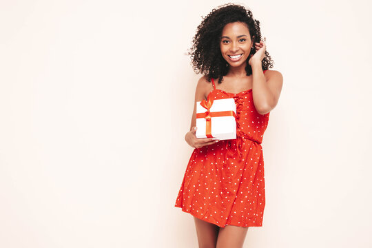 Beautiful black woman with afro curls hairstyle. Smiling model dressed in red summer dress. Sexy carefree female posing near white wall in studio. Tanned and cheerful. Holding gift box