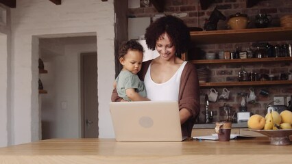 Biracial mother working from home with her young daughter in arm in a modern-styled kitchen.