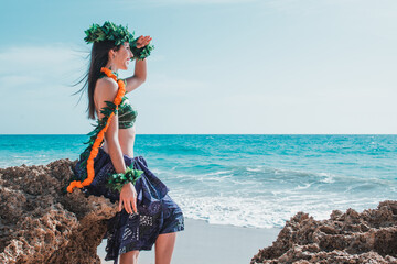Woman scanning horizon with arm raised with hand shielding sun from face wearing tropical hula dance outfit. Hawaiian woman resting relaxed on a paradisiacal beach. Oriental and exotic beauty.