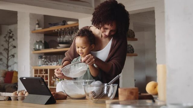 Biracial mother and daughter in modern-styled kitchen sifting flour in bowl to bake a cake.