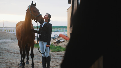 a brown horse turns its head towards the face of a young woman in front of a barn where there is another horse that cannot be seen well because it is darkened. High-quality photo